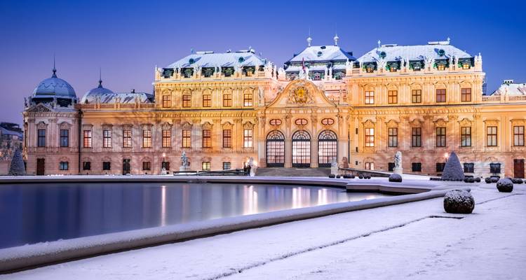 Baroque palace with a reflecting pond in a snowy setting at dusk.