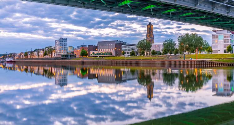 Bâtiments modernes au bord de l'eau et clocher d'église reflétés dans une rivière calme sous un pont en acier au crépuscule.