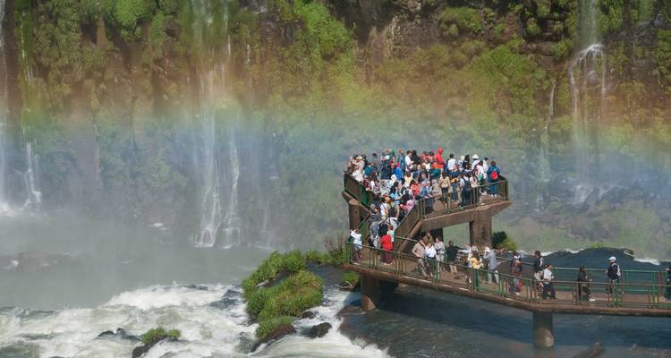 Touristen auf einer Aussichtsplattform bei den Iguazu-Wasserfällen mit einem Regenbogen.