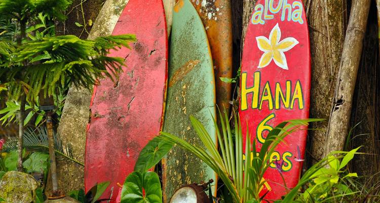 Des planches de surf aux couleurs vives et usées par le temps, ainsi qu'une pancarte peinte à la main indiquant « Aloha Hana 6 miles », encadrées par une végétation tropicale luxuriante.