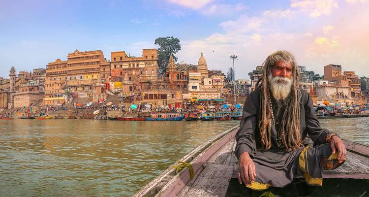 Ein älterer Mann auf einem Boot mit den Ghats von Varanasi im Hintergrund.
