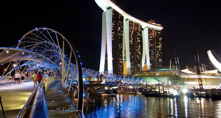 Le pont Helix illuminé mène à Marina Bay Sands, se reflétant sur les eaux calmes de la nuit avec des gens qui marchent.