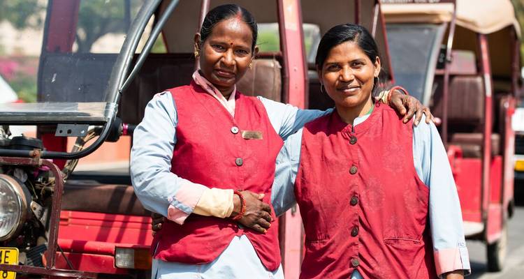 Twee vrouwen in rode vesten die voor auto-riksja's staan en glimlachen.