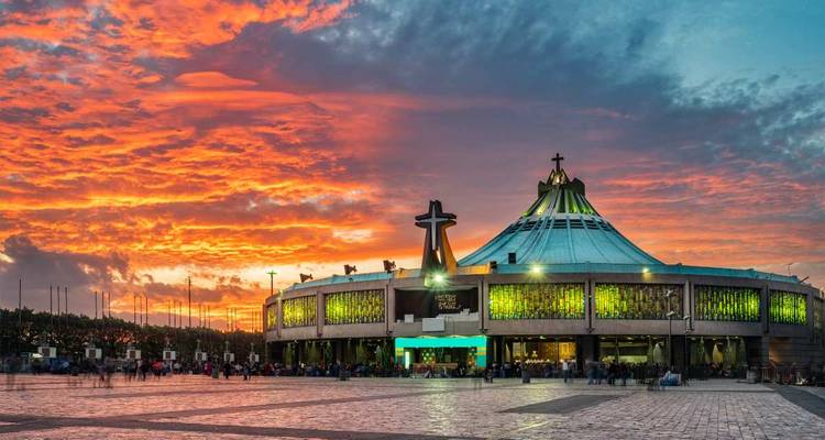 Moderne ronde basiliek met een opvallend kruis verlicht door een dramatische oranje zonsonderganghemel, menigtes verzamelen zich op het plein