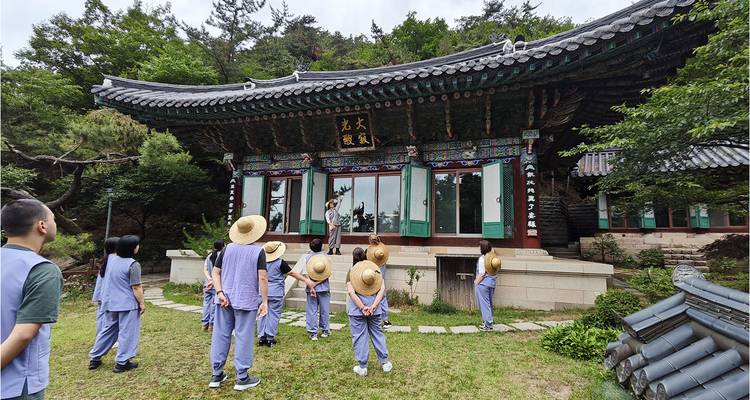 Participants in grey temple attire and straw hats stand before an ornate Korean mountain temple hall.