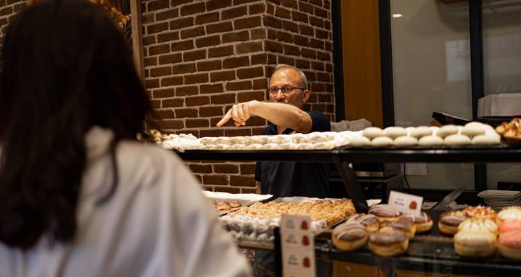 Un vendeur de boulangerie montre du doigt les pâtisseries tandis qu'un client observe à l'intérieur d'une boutique aux murs de brique.