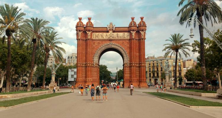 Gran arco de ladrillo en una plaza con gente y palmeras.