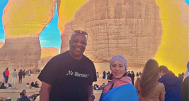 Un couple posant devant une grande arche de roche avec d'autres personnes en arrière-plan.