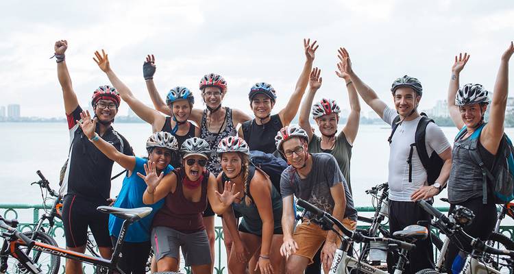 Excited group of cyclists posing by a waterfront with raised hands.