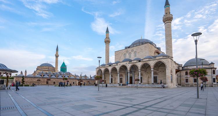 Ruim plein voor Konya's Mevlana Museum complex met torenhoge minaretten en een turquoise koepel onder een heldere hemel.