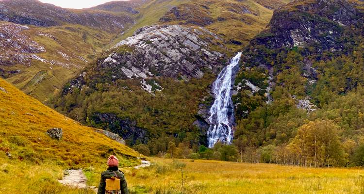 Une personne marchant vers une cascade dans une vallée pittoresque.