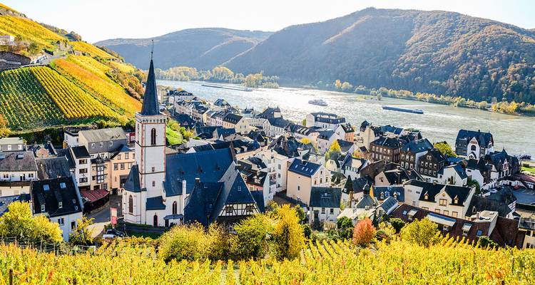 Village pittoresque de la vallée du Rhin avec clocher d'église, vignobles et péniches fluviales sous des collines boisées.
