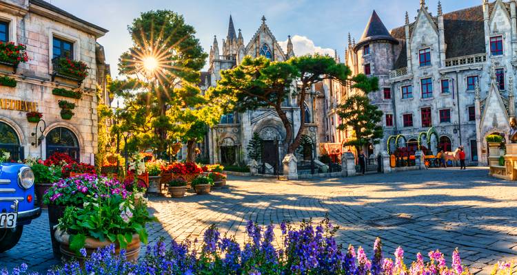 Place pittoresque avec des fleurs et des bâtiments historiques, avec la lumière du soleil filtrant à travers les arbres.