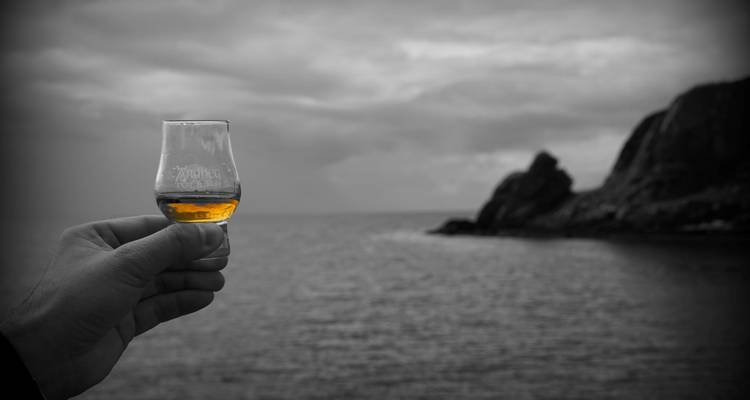 Glass of whiskey held up against a coastal landscape.