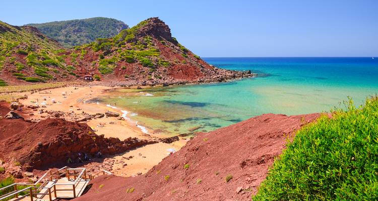 Plage de sable rouge avec collines rocheuses et eau turquoise.