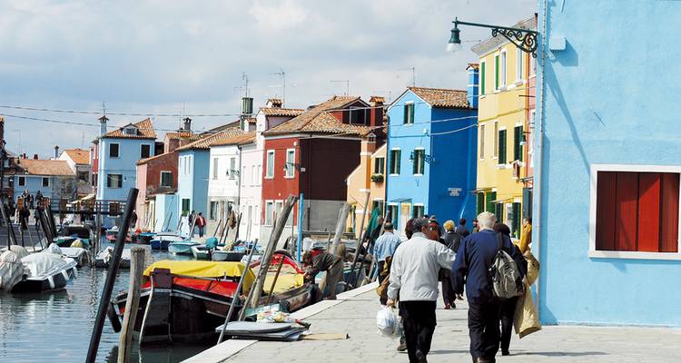 People walking along colorful houses by a canal in a small Venetian island.