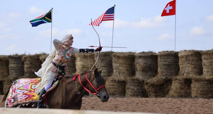 A person in traditional attire performing archery on horseback with flags in the background.