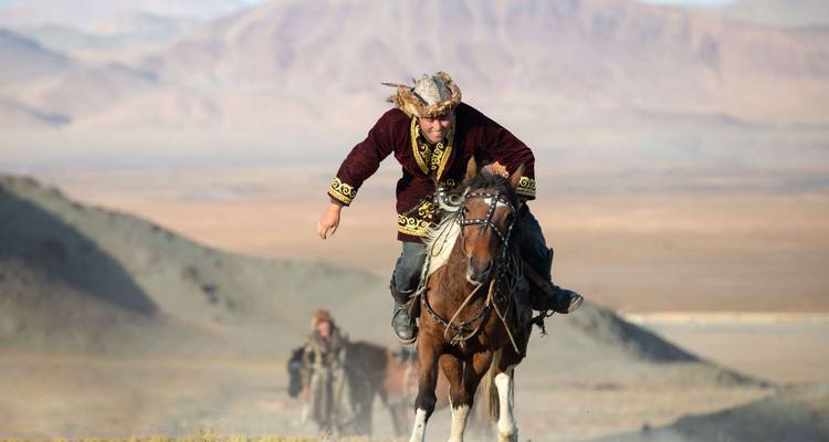 A person on horseback in traditional costume racing across mountainous terrain.