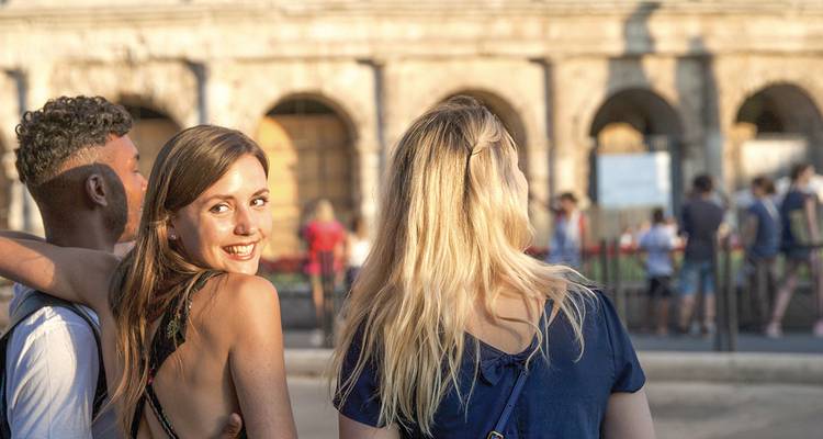 Tres amigos pasean junto a los arcos antiguos del Coliseo, uno volteando hacia atrás con una sonrisa.
