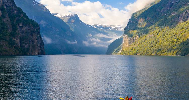 Dos kayakistas reman a través de las serenas aguas azules del Geirangerfjord de Noruega entre imponentes acantilados verdes y picos nevados