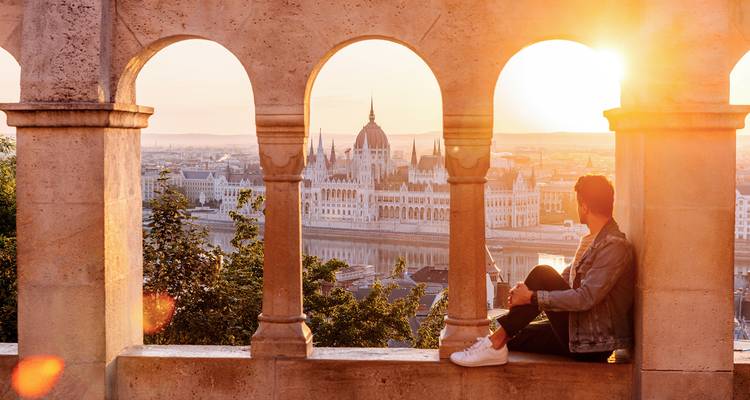 Person sitting under arches with a view of an iconic parliament building at sunset.