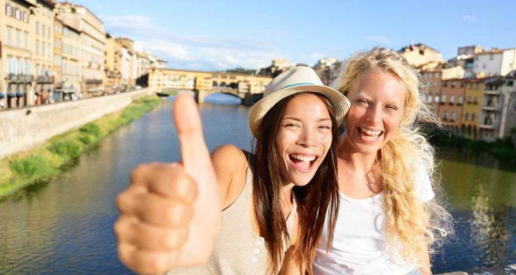 Two women smiling with a thumbs up near a river with a historic bridge.