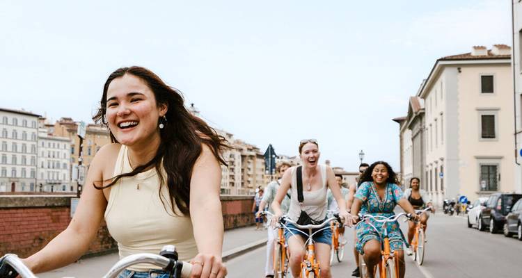 Cyclists ride through Florence on bright orange bikes alongside historic riverside buildings