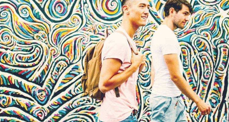 Deux jeunes hommes passent devant un mur de graffitis abstraits colorés, l'un souriant avec un sac à dos.