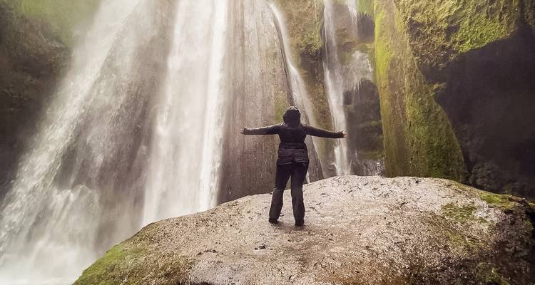 Person standing with arms outstretched in front of a waterfall.