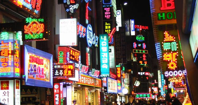 Calle bulliciosa en una ciudad asiática iluminada con neón con tiendas y letreros diversos.