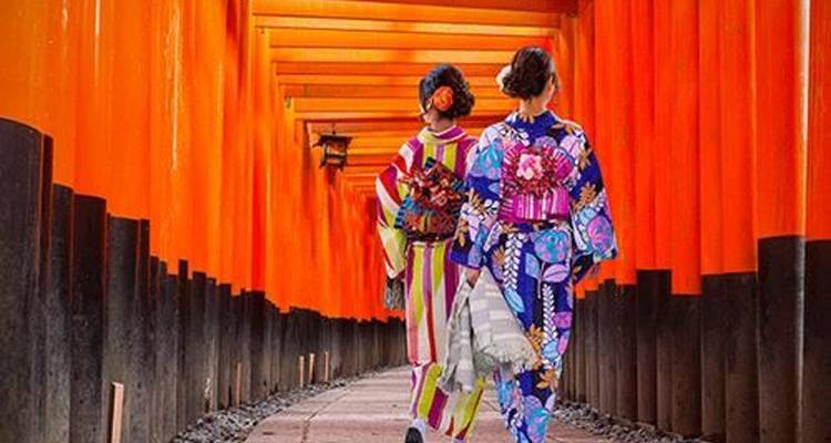 Dos mujeres en kimono caminando por un túnel de puertas torii naranjas.