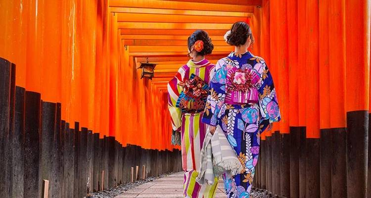 Dos mujeres en kimono caminando por un túnel de puertas torii naranjas.