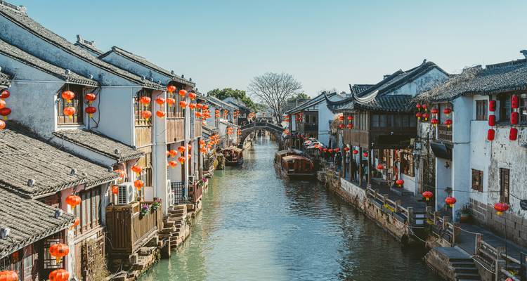 Een pittoresk kanaal omzoomd door witgekalkte Suzhou-huizen versierd met rode lantaarns onder een heldere blauwe hemel.