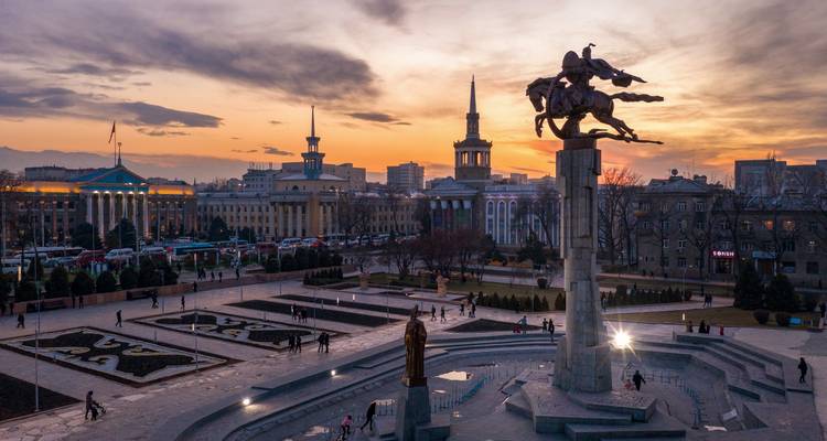 City square at sunset with a statue and several buildings in the background.