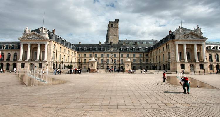 Een ruim plein met fonteiinen strekt zich uit voor het indrukwekkende Palais des Ducs in Dijon onder een gedeeltelijk bewolkte hemel.