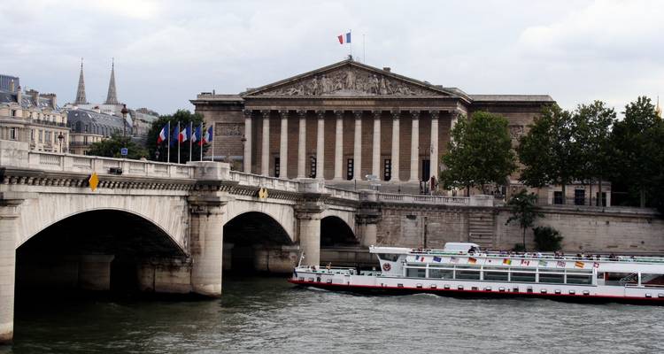 Historic building with a French flag and a bridge over a river with a sightseeing boat.