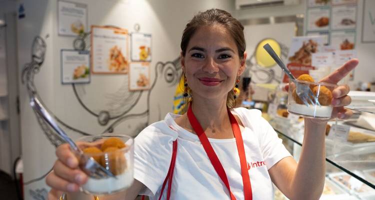 Glimlachende vrouw achter een toonbank met desserts buiten een eetgelegenheid.