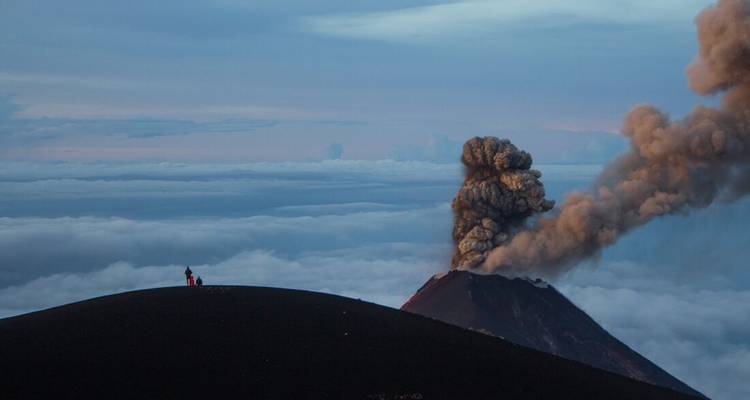Un volcan émettant de la fumée dans le ciel, vu depuis un sommet voisin au crépuscule.