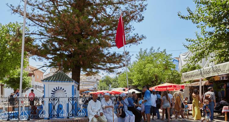 People gathered in an outdoor market under trees with some local architecture.