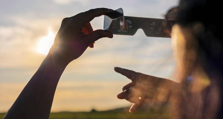 Silhouette d'une personne tenant des lunettes d'éclipse vers le soleil brillant tout en pointant vers le ciel.