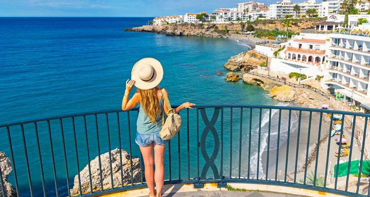 Femme portant un chapeau de paille regardant depuis un balcon la plage incurvée de Nerja et les eaux bleues de la Méditerranée.
