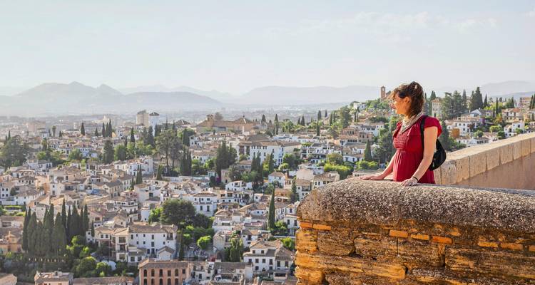 Femme vêtue d'une robe rouge se tenant près d'un mur en pierre surplombant le paysage urbain aux murs blanchis à la chaux et les montagnes lointaines de Grenade.
