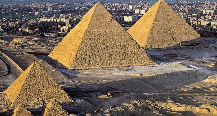 Aerial view of large pyramids in a desert with a city in the backdrop.