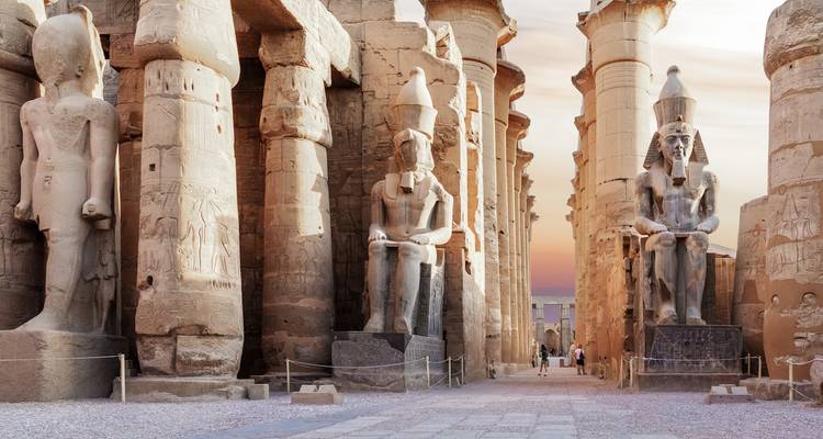 Large seated statues and columns at an ancient stone temple during sunset.