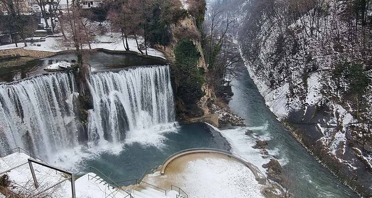 A snowy scenic view of waterfalls and a river in a forested area.