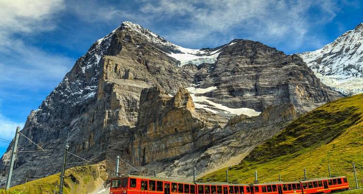 A red train set against towering rocky mountains under a blue sky.
