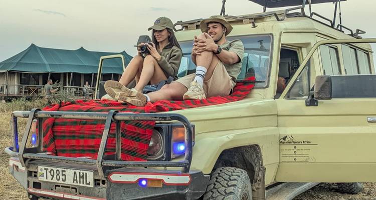Young couple sit atop a safari jeep draped in Maasai blanket at a tented camp on the Serengeti plains.