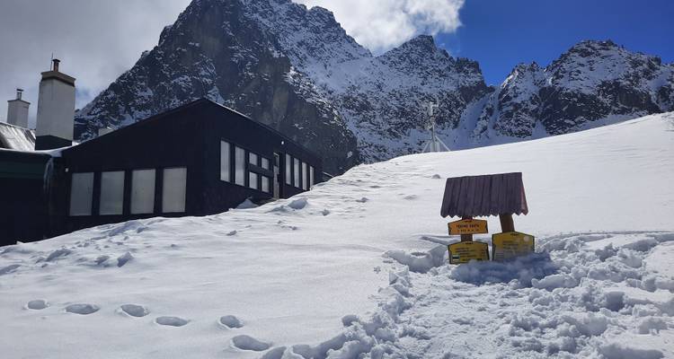 Mountain lodge partially covered in snow against a snowy peak.
