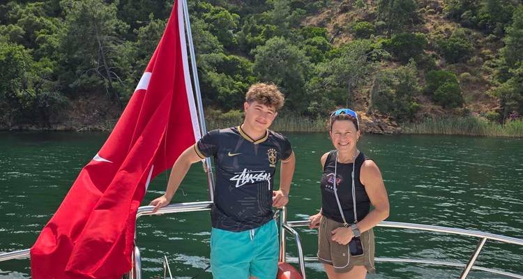 Moeder en puberseun staan op een bootdek met een Turkse vlag, met een smaragdgroen meer en met dennenbomen bedekte heuvels op de achtergrond.