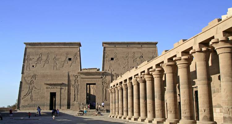 Patio de un templo antiguo egipcio con enormes pilonos y una columnata bajo un cielo azul cristalino.